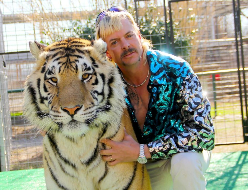 Um homem, à direita, abraça com um braço um tigre. Os dois, de frente, olham para a câmera.