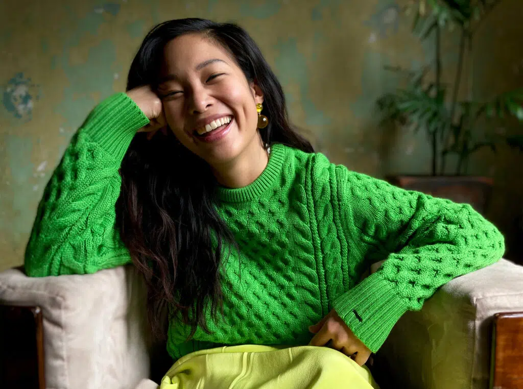 Iphone se 2020 ou iphone xr: qual o melhor modelo de entrada da apple? Foto de uma mulher asiática sorrindo tirada com um iphone se 2020. Ela veste um suéter verde de tricô claro e está sentada em uma poltrona cinza.