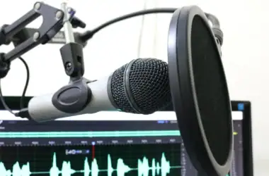 Microfone para podcasts em evidência na frente de um computador, provavelmente com algum programa de edição de áudio aberto