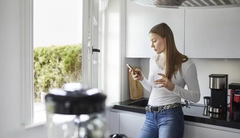 Seguro de celular? Veja opções e preços Mulher procurando por seguro para smartphone na cozinha de casa