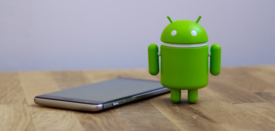Boneco do android ao lado de um smartphone, sobre uma mesa de madeira e um fundo cinza