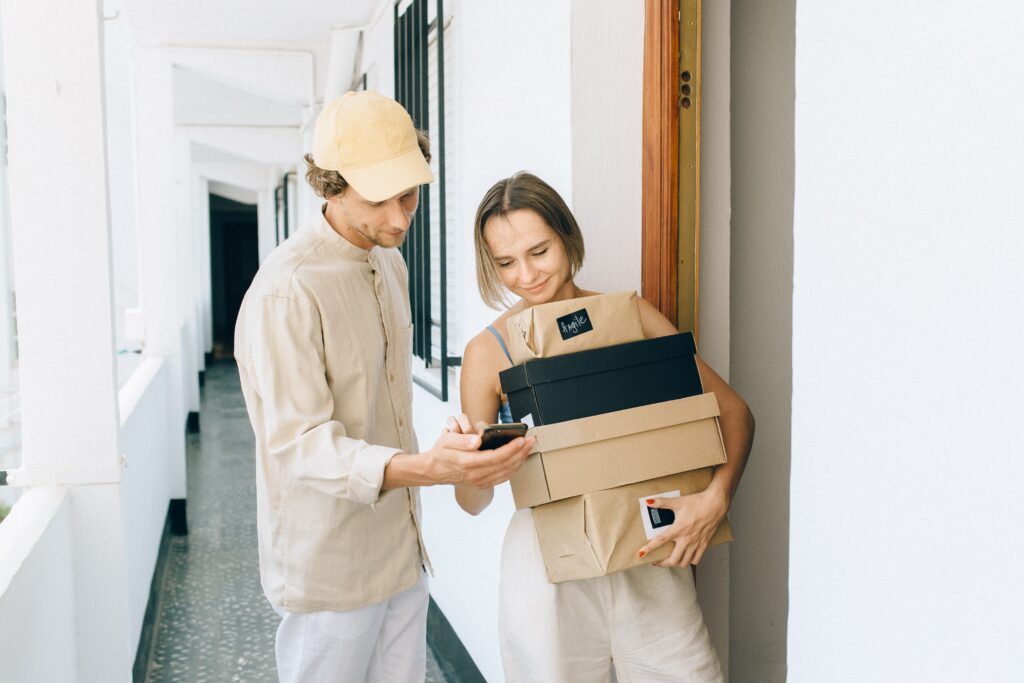 A imagem mostra um entregador com celular na mão e uma mulher com pacotes de encomendas recebidos.