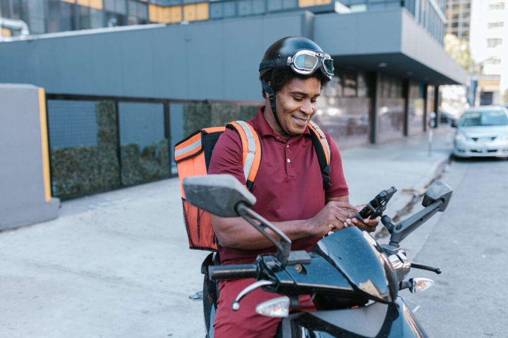A imagem mostra um entregador do mercado livre parado em cima da moto com mochila nas costas e um celular na mão.