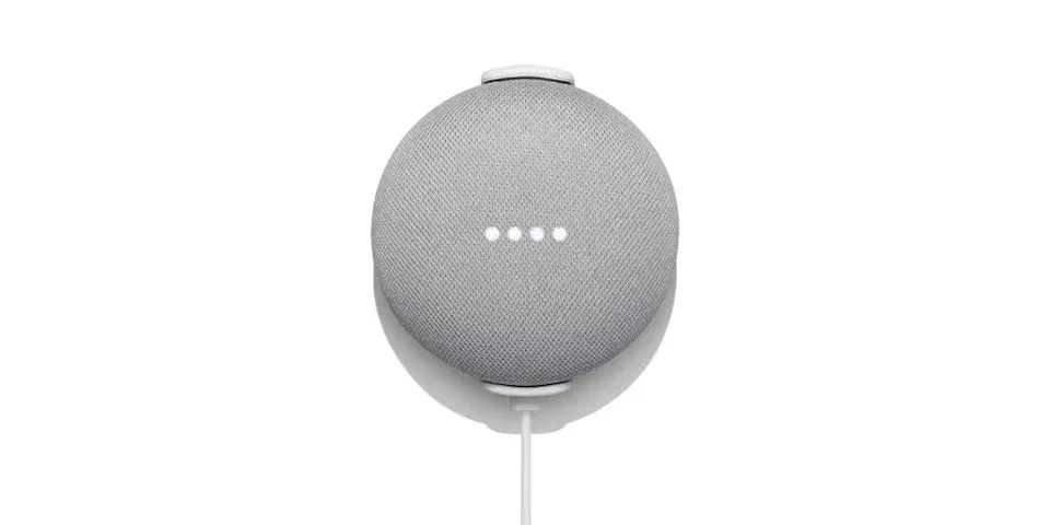 Qual o custo de uma casa conectada do google home? Parede branca e nest hub mini no meio na cor cinza, 4 pontos de luz no meio indicando que está ligado e um cabo de energia conectado no aparelho.