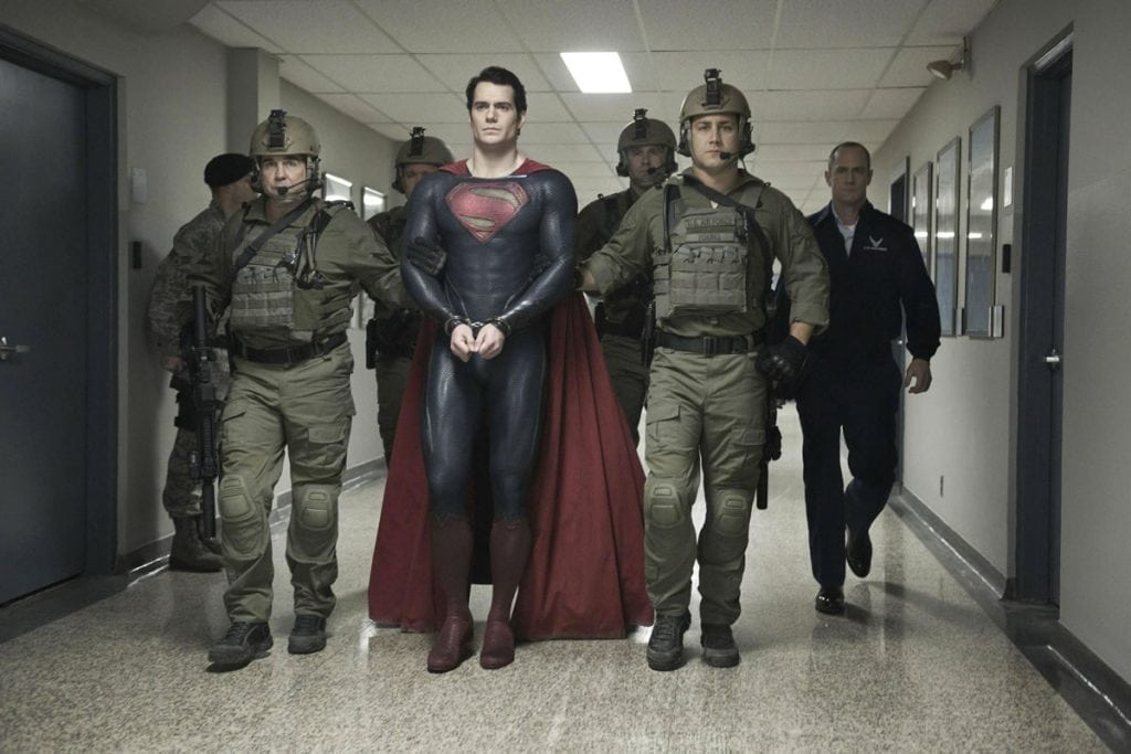 Alt: superman vestido como herói em uniforme preto com capa vermelha, cercado por soldados em uniforme tático.