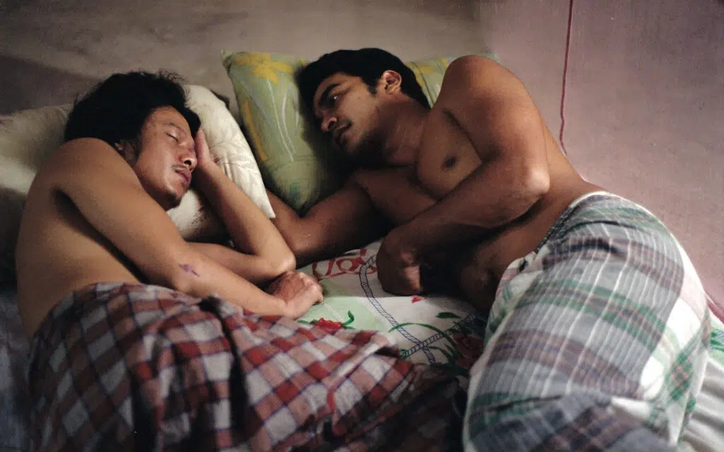 Imagem de dois homens nus, deitados na cama e trocando olhares carinhosos, representando temática lgbtqiap+.