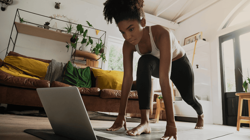 Imagen de una mujer realizando ejercicios de estiramiento o calentamiento en casa, usando una computadora portátil como guía.