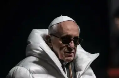 Imagem de um idoso com óculos escuros, vestindo jaqueta branca, com fundo escuro.