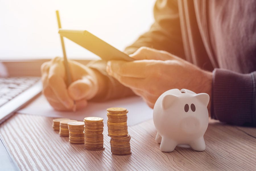 Compras internacionais online valem a pena? Um guia completo Na foto um homem faz cálculos com um lápis, a sua frente vê-se moedas empilhadas e um cofre de porcelana em formato de porquinho.