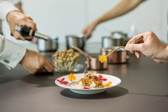 Experiência gastronômica t alt 20 prato com cores amarelo e vermelho, em mesa de cor marrom escuro, com mãos de cozinheiro em volta, em segundo plano