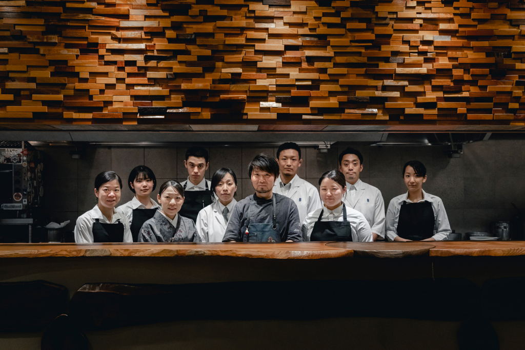 Menu do restaurante 21 talt cozinheiros vestindo roupas de cores escuras e brancas, atrás de uma bancada; ao fundo, parede amadeirada, com textura em relevo