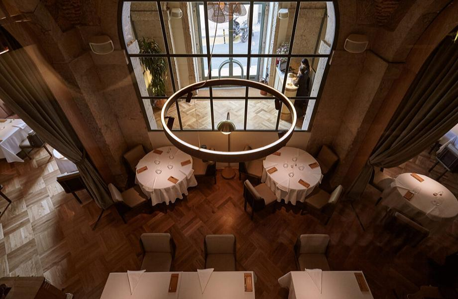 Menu do restaurante t alt 25 mesas de restaurante vistas de cima, em sala de tom amadeirado, com janela ao centro, que está por trás de um lustre vazado, composto por vigas finas