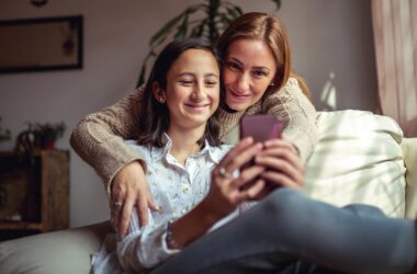 Mulher e menina sorrindo juntas enquanto olham celular na sala de estar.