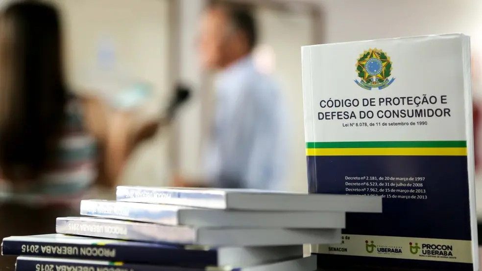 Produto há mais de 30 dias no conserto? Você pode receber um novo, segundo o cdc Em primeiro plano, na foto, há uma pilha de livros do código de defesa do consumidor. Em segundo plano e desfocado, há uma pessoa ao fundo, aparentemente sendo entrevistada.