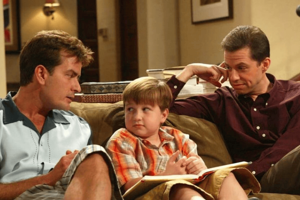 Voor x na: kijk hoe kindacteurs er vandaag de dag uitzien! Een jongen zit op een bank tussen twee pratende volwassenen, een scène uit een familieserie of -film.