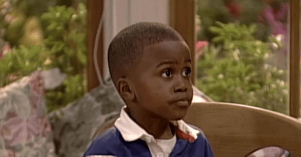 Antes x depois: veja como estão atores mirins hoje! Menino afrodescendente com cabelo raspado, expressão pensativa, sentado em ambiente doméstico, cenário com janelas e plantas.