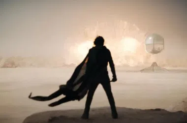 Relógio futurista no deserto com explosão ao fundo, cenário de ficção científica e tecnologia avançada.