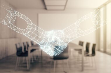 A imagem é um holograma de duas mãos se apartando como se estivessem fazendo um trato. Ao fundo é possível ver uma sala de reuniões.