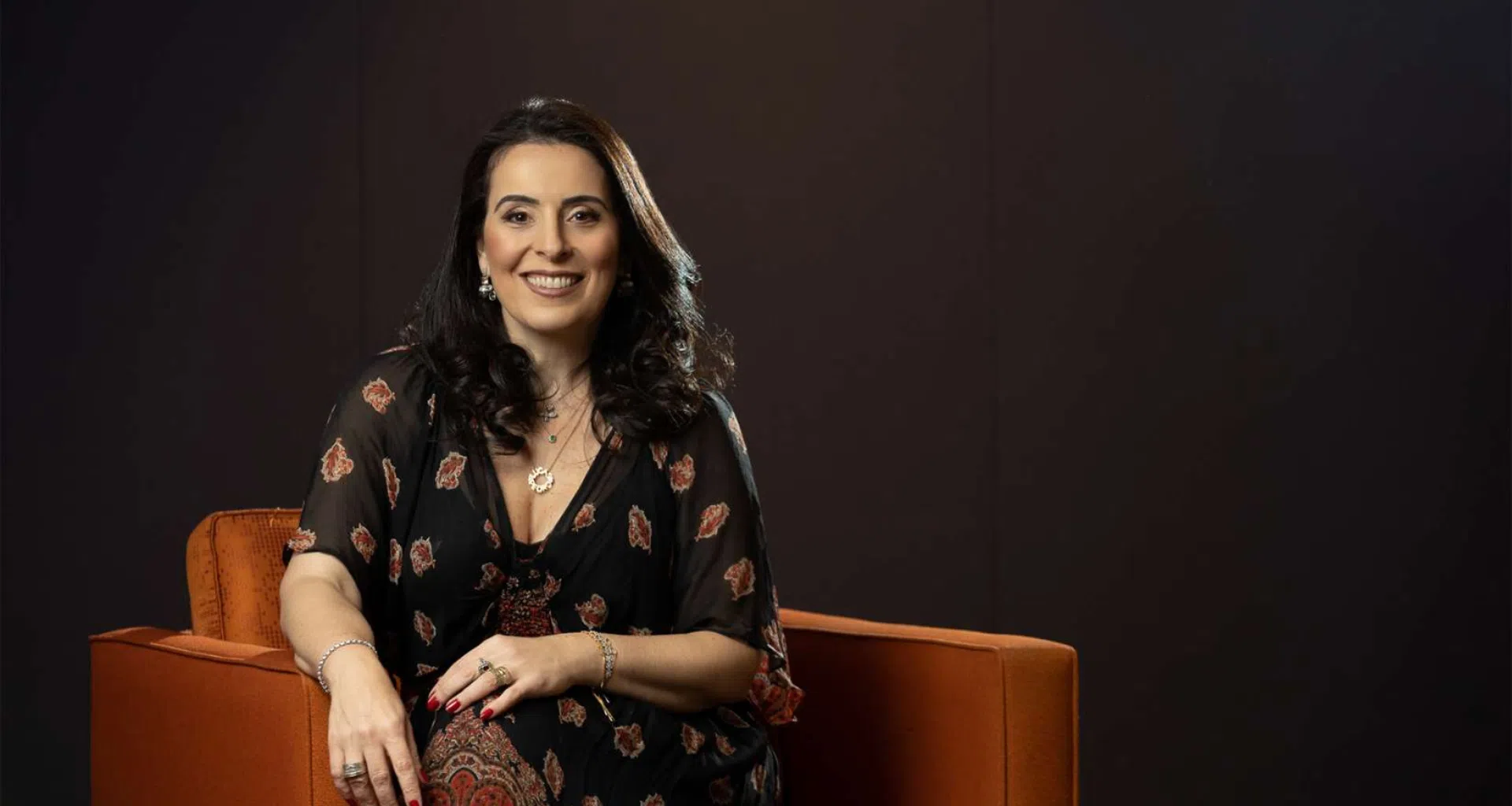 Mulher sorrindo sentada em sofá laranja, estilo profissional, em ambiente de estúdio.