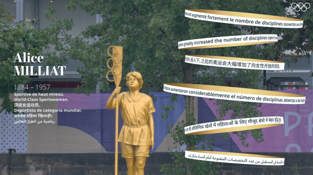 At alt text: estátua dourada de alice milliat, pioneira do esporte feminino, na rua com árvores ao fundo.