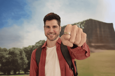 Homem sorridente apontando para a câmera com monumento ao fundo, promovendo viagens e turismo cultural.