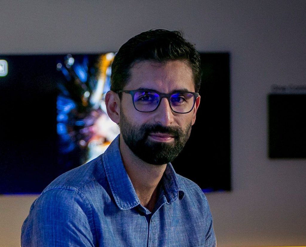 1. Homem com óculos escuros e barba em ambiente com tv ao fundo.