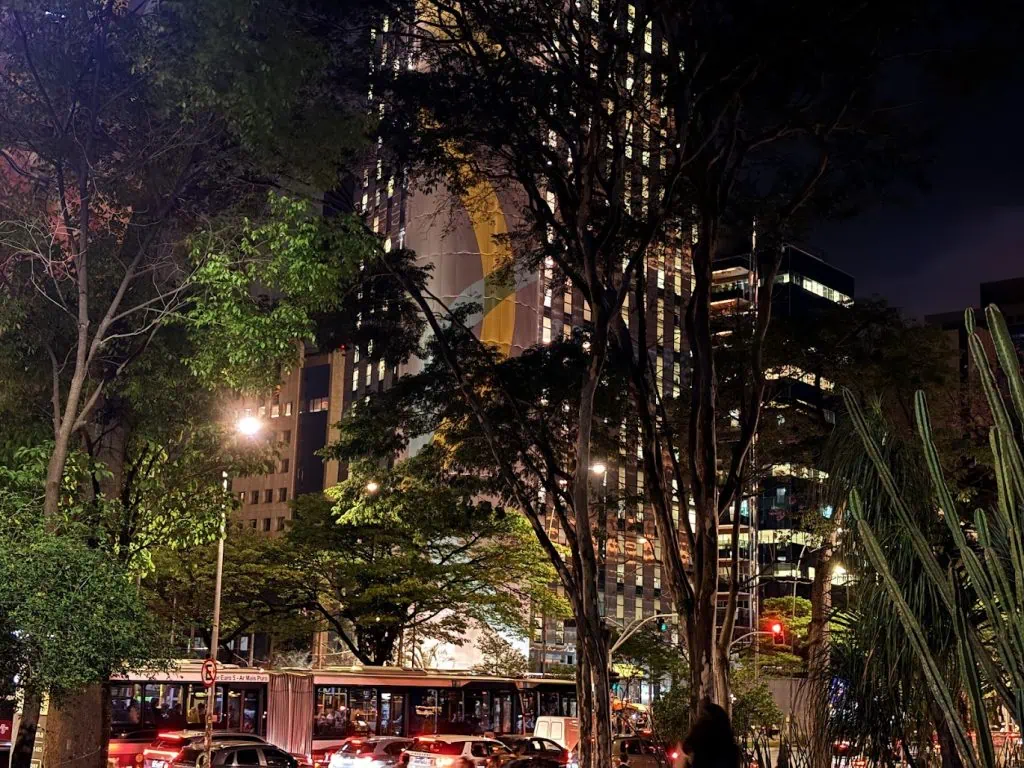 Anmeldelse: iPhone 16, hjemmehørende i en tidsalder av kunstig intelligens Foto av en moderne bygning opplyst om natten i São Paulo, med trær foran og tett trafikk.
