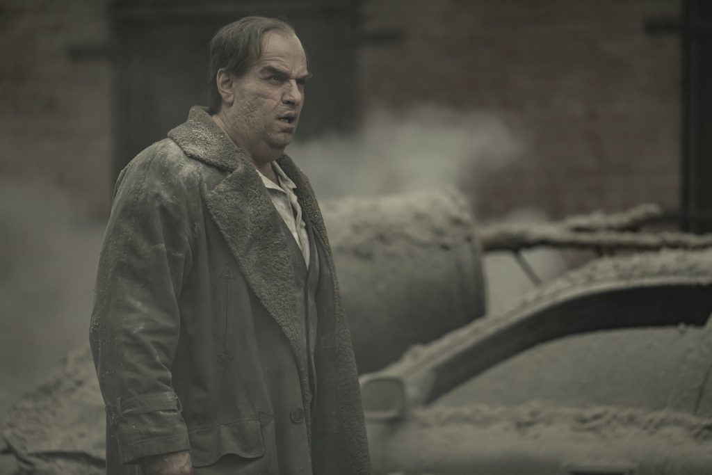 Homem em situação de desastre com fumaça ao fundo em cenário urbano destruidor.