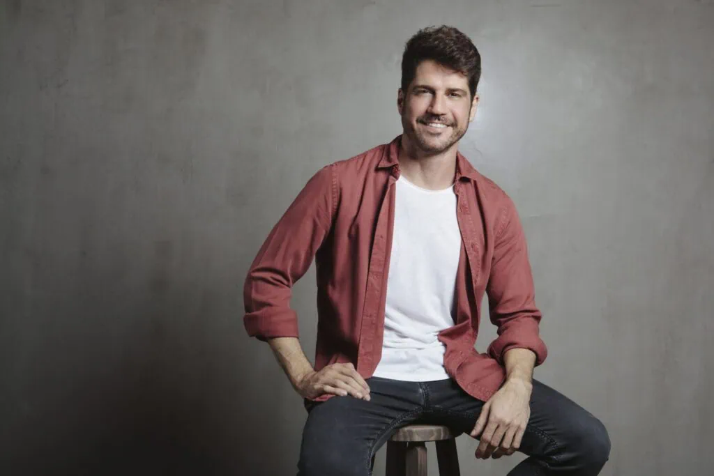 Contato Homem sorridente sentado em banquinho, estilo casual, fundo neutro, atenção para a expressão amistosa.