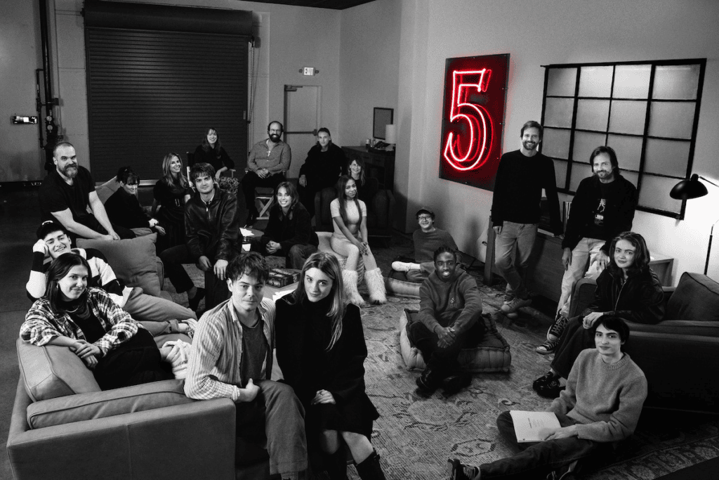 Elenco e produção de stranger things em uma sala, foto em preto e branco, exceto o número  na parede com tons de vermelho