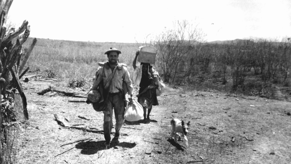 Homens caminhando em trilha desértica com animais e bagagem, cenário de perseguição ou migração.