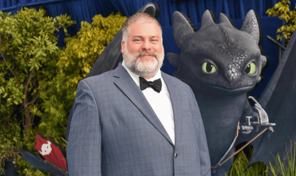Homem com barbae terno em evento de estreia de 'como treinar seu dragão' com personagem do filme ao fundo.