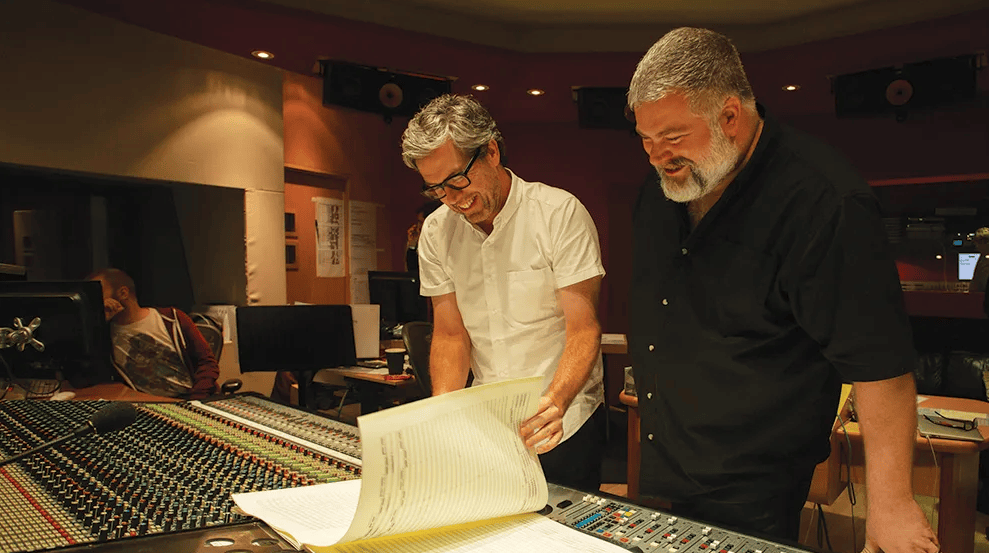 Estúdio de gravação com duas pessoas analisando papéis e equipamentos de áudio.
