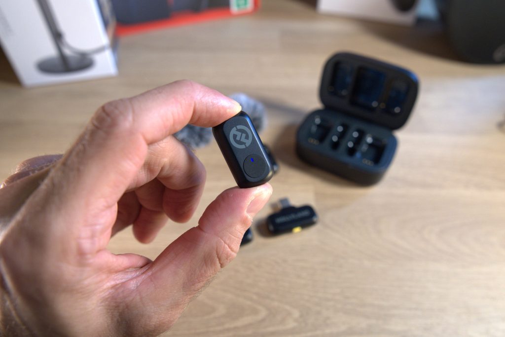 Review: hollyland lark a1, o kit de microfones sem fio com melhor custo-benefício de 2025 A imagem mostra uma mão bem de perto segurando o microfone e ao fundo desfocado, podemos percebes os acessórios do lark a1