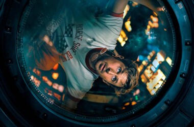 Imagem de um jovem olhando através de uma máquina de lavar roupa, com luzes de cidade ao fundo.