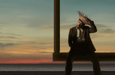 Imagem de bad bunny sentado em um balanço na praia ao pôr do sol, usando chapéu de palha e traje cas.