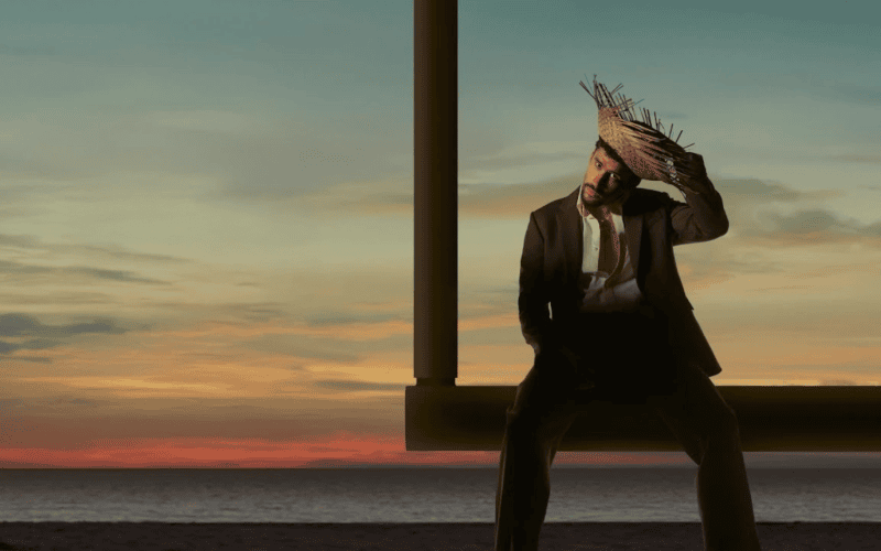 Imagem de bad bunny sentado em um balanço na praia ao pôr do sol, usando chapéu de palha e traje cas.