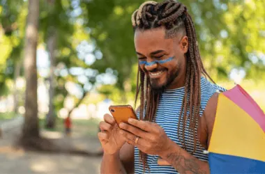 Jovem com celular, sorrindo, em parque com guarda-chuva colorido ao lado.