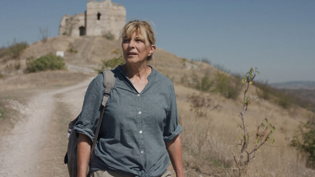 Cena de uma mulher de blusa de botão azul e uma mochila caminhando em um deserto durante o dia
