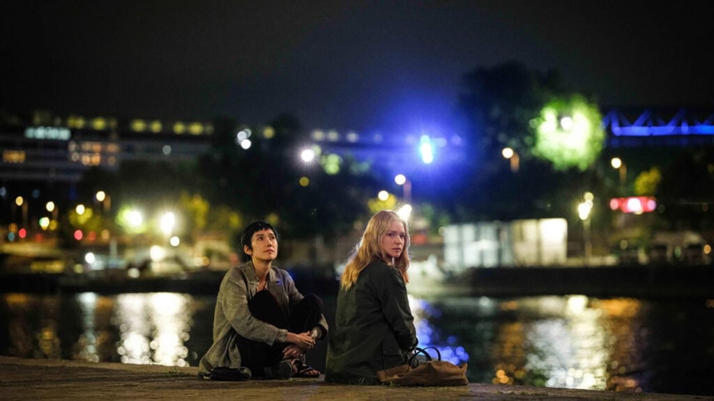 Cena de duas mulheres à beira de um rio em uma cidade no período da noite