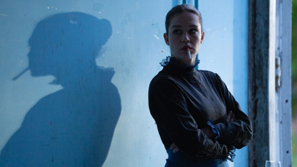 Cena de uma mulher de blusa azul e braços cruzados e sua sombra projetada à esquerda em uma parede azul clara