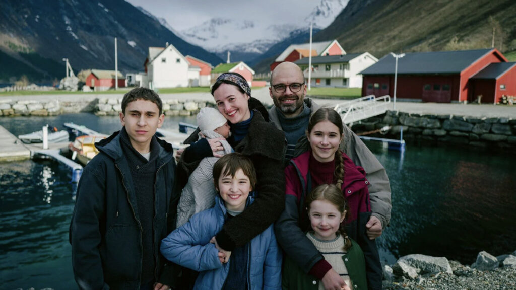 Cena de uma família de 6 pessoas sorrindo. Ao fundo várias casas sob um lago