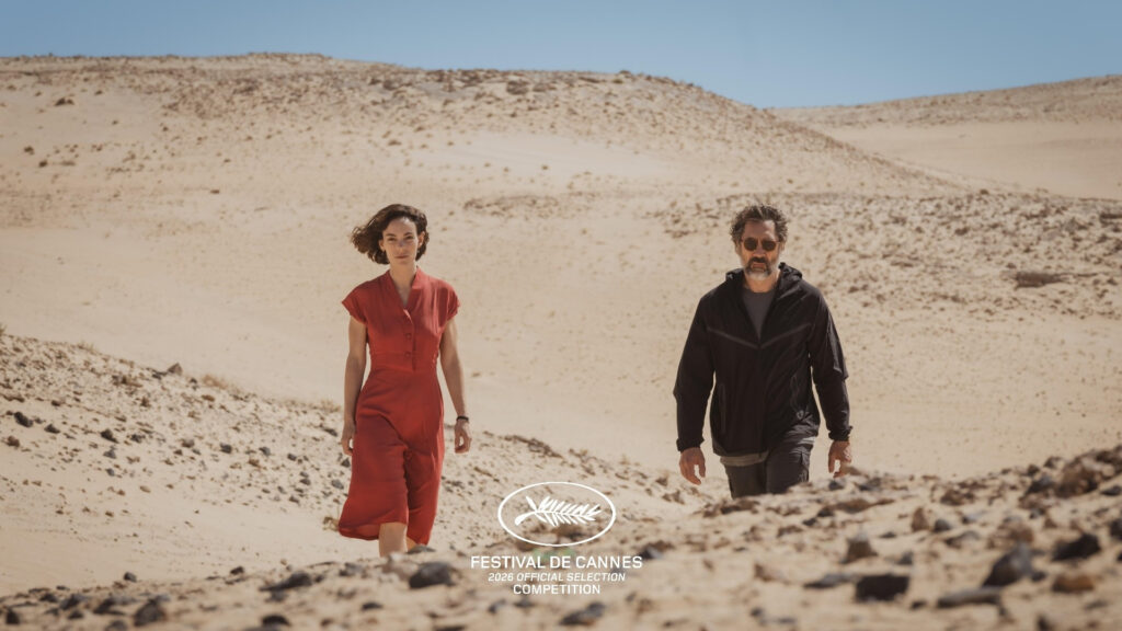 Cena de uma mulher de vestido vermelho e um homem de moletom preto caminhando de um deserto