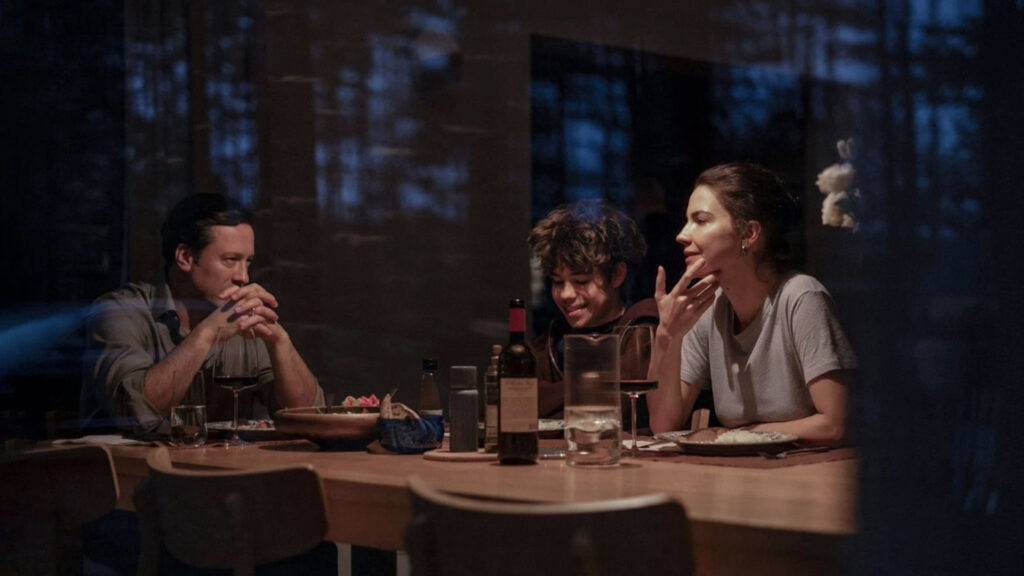 Cena de um homem sentado de mãos cruzadas olhando atentamente a uma mulher e uma garota do outro lado da mesa