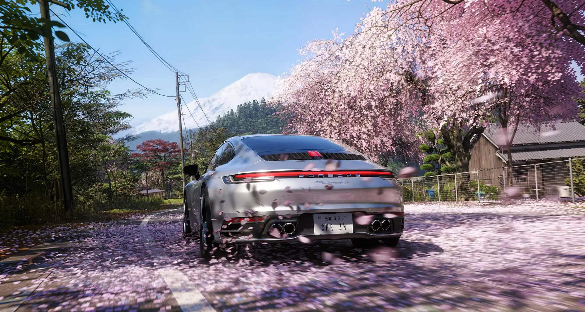 Porsche prata dirigindo em estrada rural japonesa coberta por pétalas de cerejeira rosa, com o monte fuji nevado ao fundo e árvores de sakura em flor à direita da via sob céu azu