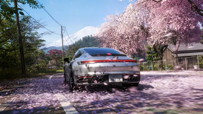 Porsche prata dirigindo em estrada rural japonesa coberta por pétalas de cerejeira rosa, com o monte fuji nevado ao fundo e árvores de sakura em flor à direita da via sob céu azu