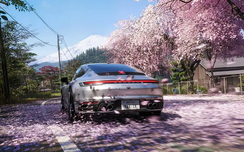 Porsche prata dirigindo em estrada rural japonesa coberta por pétalas de cerejeira rosa, com o monte fuji nevado ao fundo e árvores de sakura em flor à direita da via sob céu azu