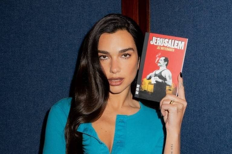 Dua lipa segura o livro jerusalem diante de fundo azul em retrato vertical.