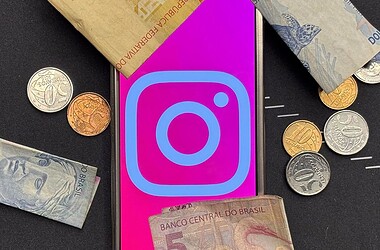 Celular com o logo do instagram cercado por cédulas e moedas de real brasileiro, representando a assinatura paga instagram plus.