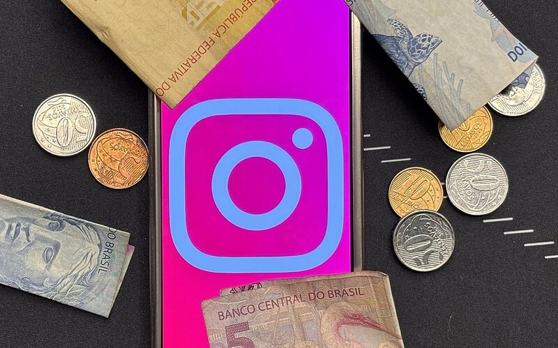 Celular com o logo do instagram cercado por cédulas e moedas de real brasileiro, representando a assinatura paga instagram plus.